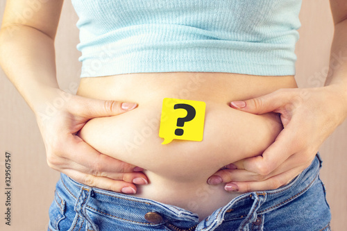 A yellow paper blank message is written question mark on belly with hands squeezes at the waist of a young woman in jeans. The concept of excess weight, weight loss, diet, obesity, junk food.
