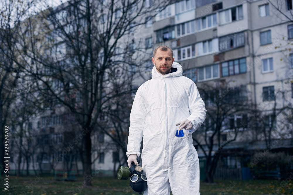 Portrait of a male scientist in white coverall, holding gas mask in one hand and glass flask in other, standing in front of a high residential building after examining the territory Portrait of a male scientist in white coverall, holding gas mask in one hand and glass flask in other, standing in front of a high residential building after examining the territory