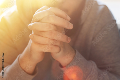 Bearded adult man praying to God sitting at home in the sunbeam. A Muslim or Christian raises his hands to God. Crossed hands in prayer gesture close up