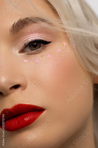 Half close-up portrait of a woman with shiny clean skin and curly blond hair. Rhinestones stars on the eyes  evening makeup and red lipstick. soft care  full lips  long eyelashes and thick eyebrows