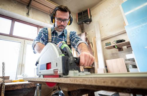 Woodworker wearing safety gear using a circular saw to cut a wood at woodwork...