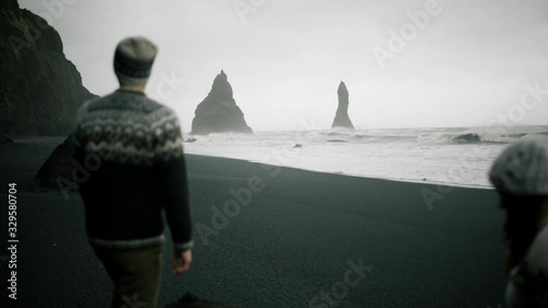 Wide angle shot of a young couple dressed in traditional icelandic sweater pointing with a finger to mountains on a black beach near Vik in Iceland photo