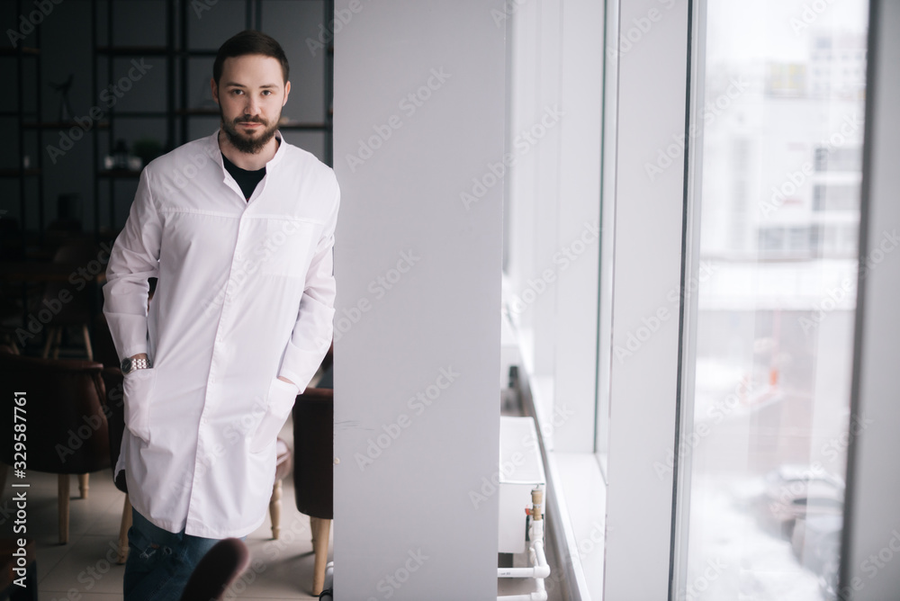 Serious doctor with a beard stands by the window and looking at camera. Male doctor posing in modern meeting office room of the hospital. Concept of medical work. Serious doctor with a beard stands by the window and looking at camera. Male doctor posing in modern meeting office room of the hospital. Concept of medical work.