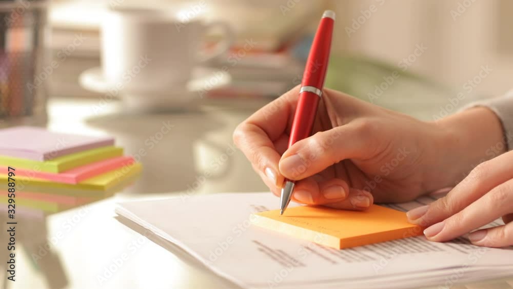 Close up of a woman hands writing reminder on orange stiky note on a desk at home