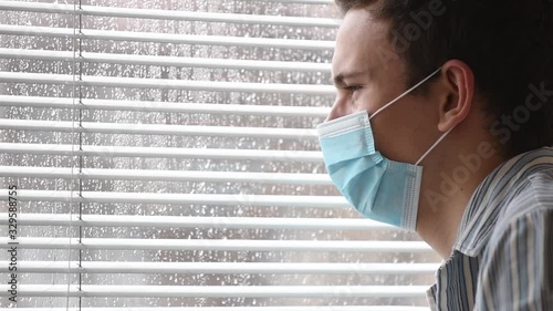 Young man wearing a face mask, sick with COVID-19, quaranteened in a hospital, looking through blinds