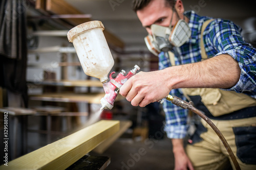 Carpenter with paint spray gun painting wooden plank in carpentry workshop
