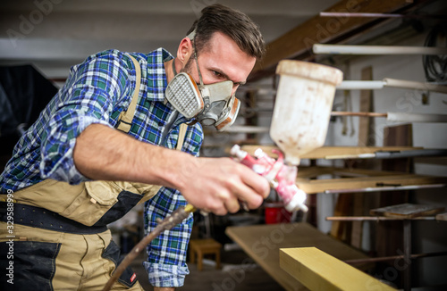 Carpenter with mask using paint spray gun to painting wooden plank in carpent...