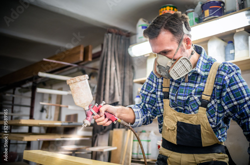 Carpenter with mask using paint spray gun to painting wooden plank in carpent...