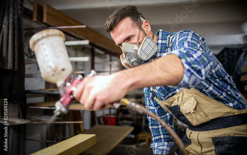 Carpenter with mask using paint spray gun to painting wooden plank in carpent...