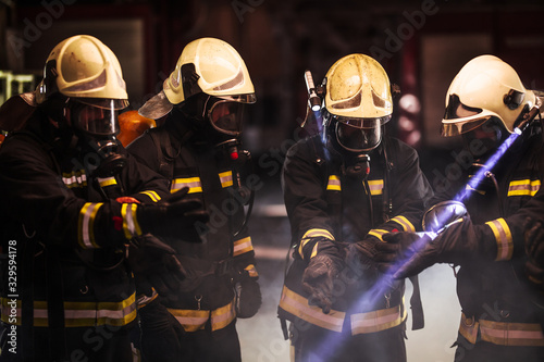 Group of professional firefighters. Firemen wearing uniforms, protective helmets and oxygen masks. Smoke in the background. © Nikola Spasenoski