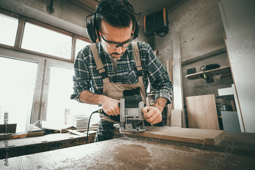 Carpenter holds milling machine and and working at wooden board in woodworkin...