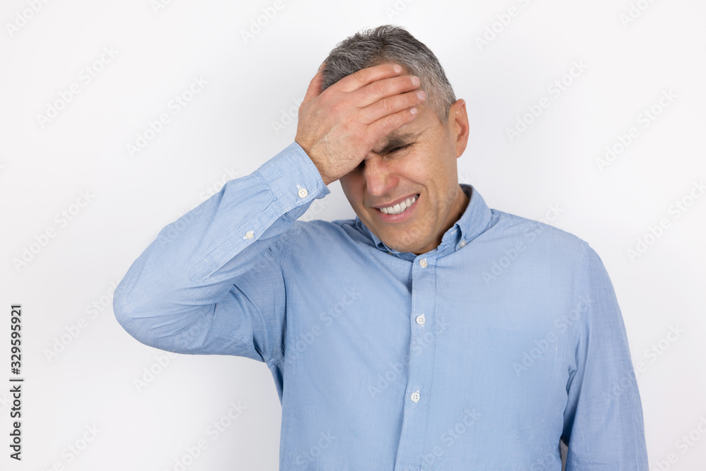 adult handsome man with grey hair wearing blue shirt touching forehead with hand feeling sorry standing on isolated white background adult handsome man with grey hair wearing blue shirt touching forehead with hand feeling sorry standing on isolated white background