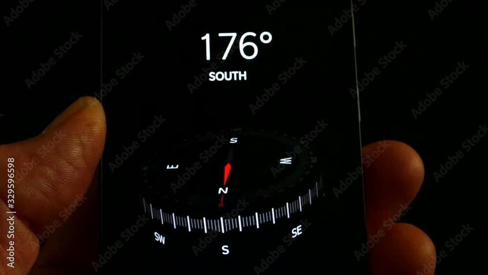 Fingers holding a mobile phone with a compass app displayed, showing the north, south, east, west direction pointer and the geographic decimal degrees as the compass moves.