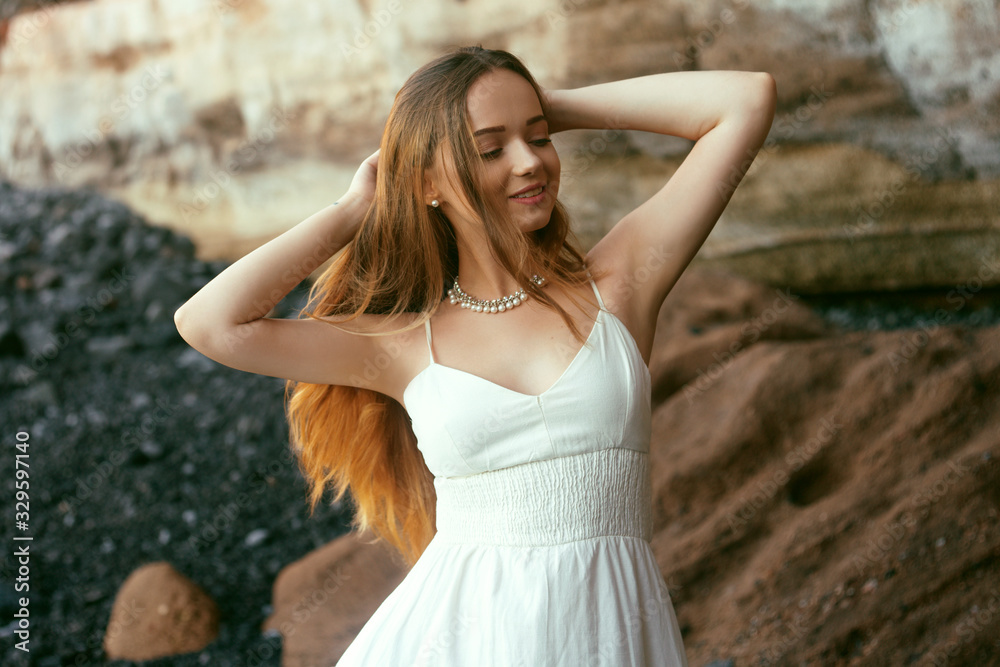 Gorgeous girl in a long white dress on the ocean at the beach. The model is inherited by nature. Girl on vacation, light dress, summer, fashion. Gorgeous long hair develops in the wind. Tenerife