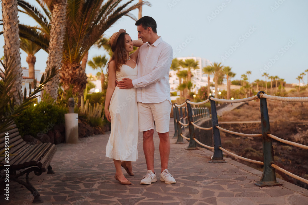 A couple strolling along the coast near the ocean on a background of palm trees. Girl in a long white dress with a man in a white date. Honeymoons, travel, tourism, Canary Islands.