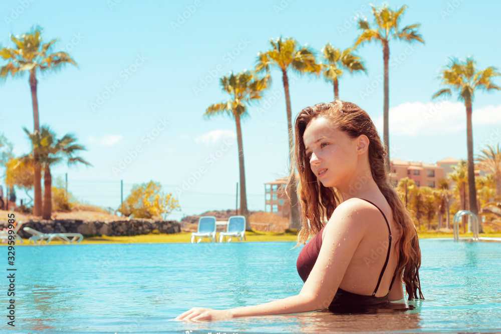 Happy girl in the pool, blonde with long hair in a red bathing suit. The girl rejoices in traveling, flirts, smiles, rejoices. Traveling. Girl on a background of palm trees.