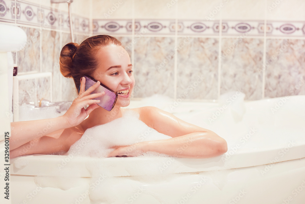 Obraz premium The girl is lying in the whirlpool with foam and talking on a waterproof phone. The girl relaxes and gossips. Poetz girl in the foam. Conversation with a girlfriend.