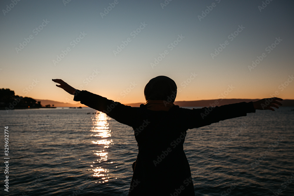 photo of young European Muslim women with hijab standing and looking into sea. She is standing turned back with her hands in the air. She is happy and relaxed. Photos taken during sunrise. 	