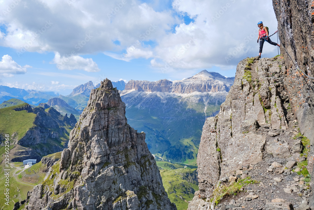 Woman on via ferrata Delle Trincee (meaning Way of the trenches), high above impressive pointy rock peak, with Sella group in the distance as background. Adventure activity in Dolomites, Italy.