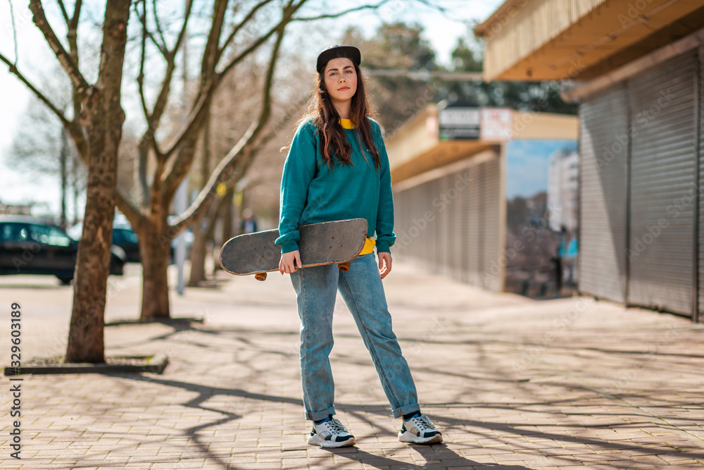 A young caucasian hipster woman stands posing with a skateboard in her hands. In the background, an alley. Concept of sports lifestyle and street culture