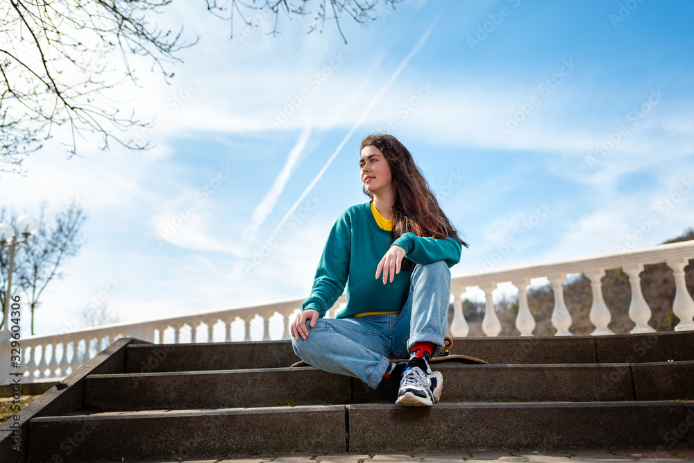 Obraz premium Caucasian teenage girl sitting on a skateboard and looking into the distance.In the background, the blue sky and the Boulevard.Bottom view.Concept of sports lifestyle and street culture