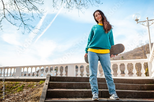 A young Caucasian woman holding a skateboard behind her back is standing on the steps. In the background, blue sky and trees. Bottom view. Copy space. Concept of sports lifestyle and street culture