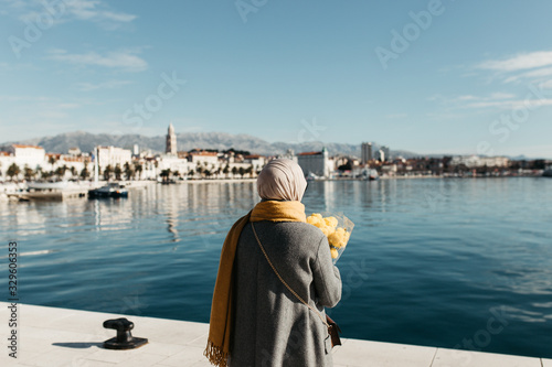 portrait of European Muslim women with hijab sitting on the stone beach with sea and port in the background. She is holding bouquet of yellow flowers. © Melika