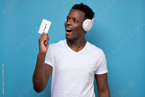 African american young man in headphones holding an old music tape. I do not understand how to use it. Studio shot on blue wall. Old retro technology concept