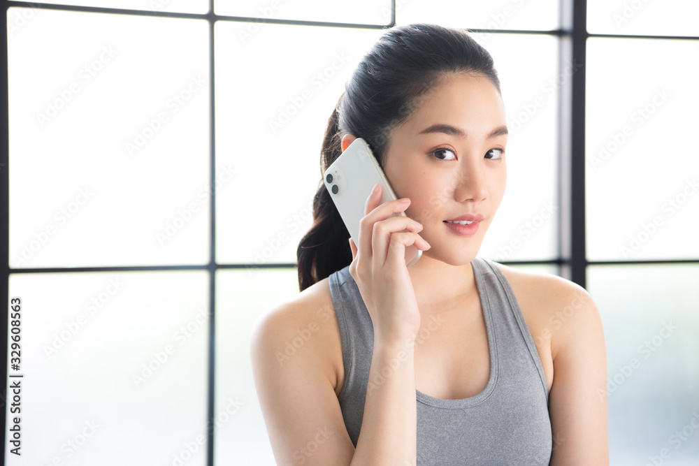 Young and beautiful asian girl wearing sportwear calling someone with her stylish smartphone in front of the window.