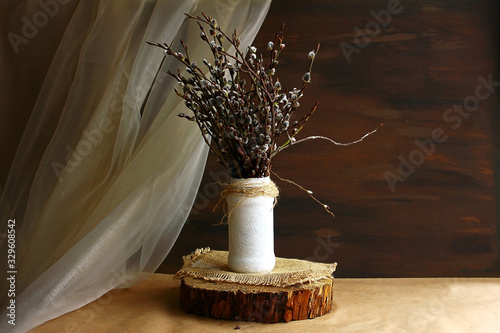 Willow branches in a white vase on a wooden stand cut, chiffon beige golden tulle on a dark brown background. The interior is in a rustic pastoral eco style.