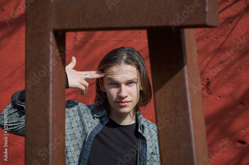 Sarcastic young man pointing his fingers to his head symbolising a pistol ready to pull the trigger, in front of a rusty red metal background.