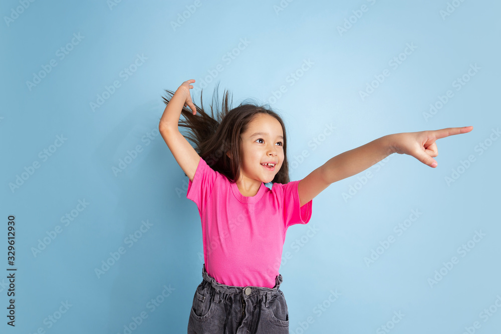 Pointing on in jump. Caucasian little girl's portrait on blue studio background. Beautiful female model in pink shirt. Concept of human emotions, facial expression, sales, ad, youth, childhood.