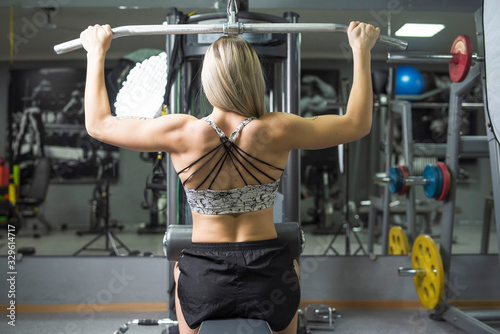 Blonde girl is engaged on a simulator in a fitness room. Photographed from behind.