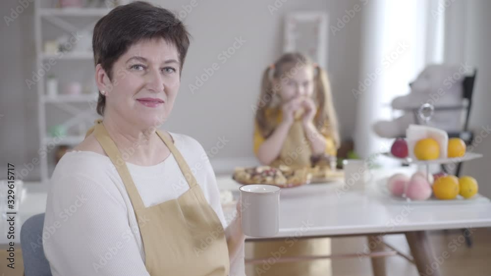 Positive brunette Caucasian woman drinking tea and smiling at camera as blurred little girl eating pancakes at the background. Happy grandmother spending Shrove Tuesday with granddaughter. Positive brunette Caucasian woman drinking tea and smiling at camera as blurred little girl eating pancakes at the background. Happy grandmother spending Shrove Tuesday with granddaughter.