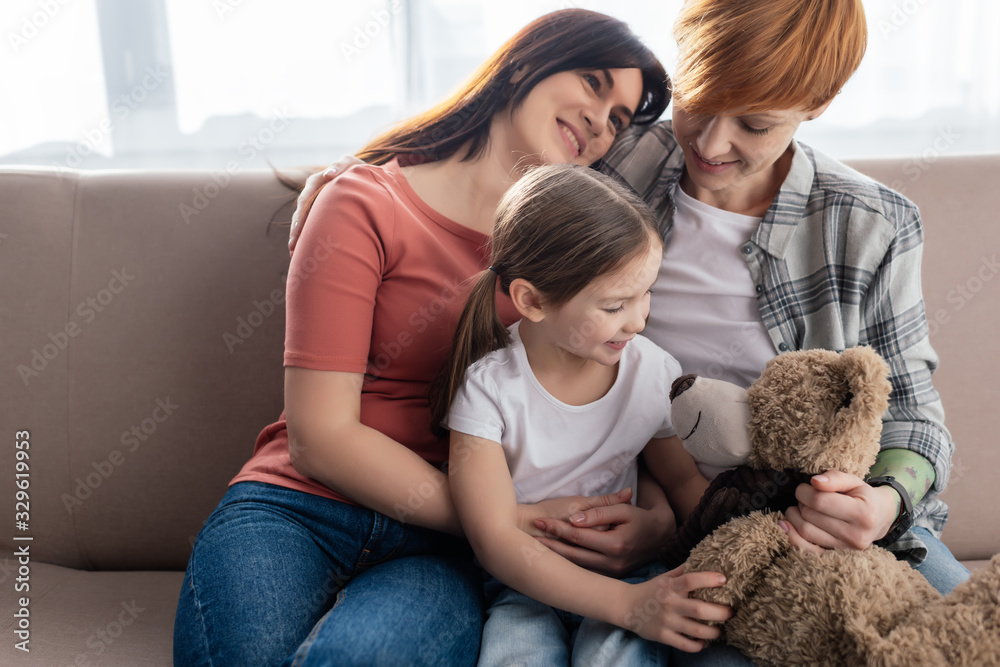 Smiling same sex parents sitting near daughter with teddy bear on couch Smiling same sex parents sitting near daughter with teddy bear on couch