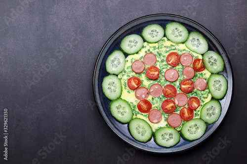 Omelet with cherry tomatoes green onions and sausages in a plate on a dark background. Top view, flat lay