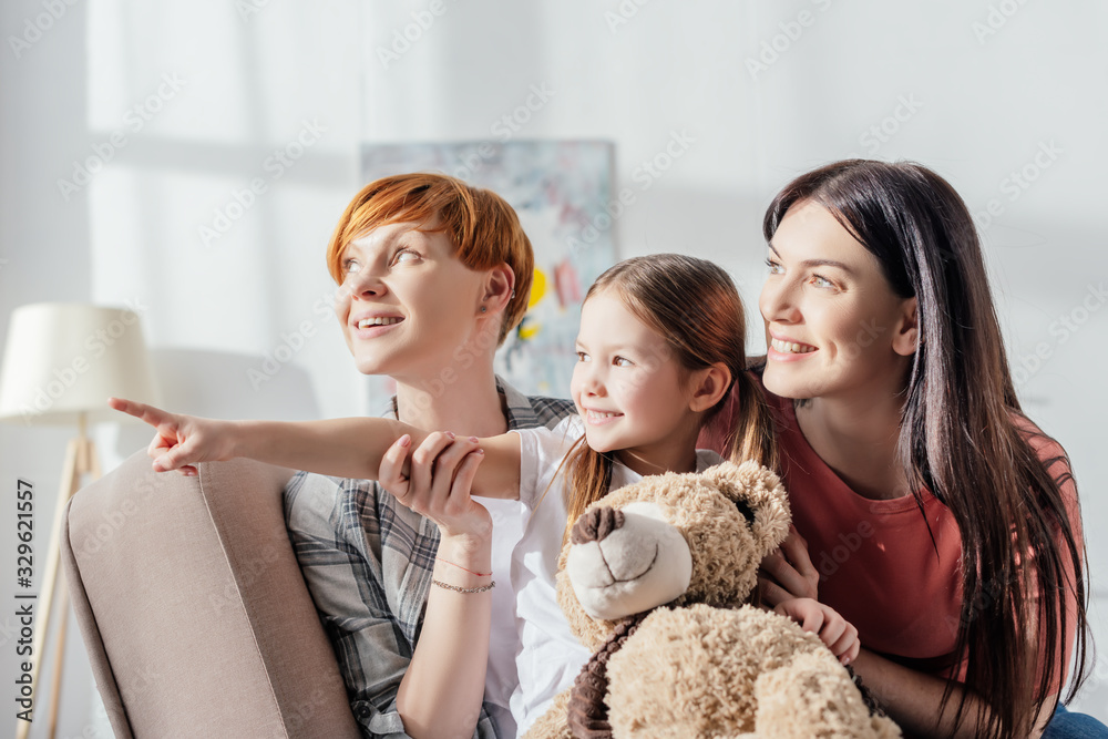 Smiling kid with teddy bear pointing with finger near mothers while sitting on couch in living room Smiling kid with teddy bear pointing with finger near mothers while sitting on couch in living room