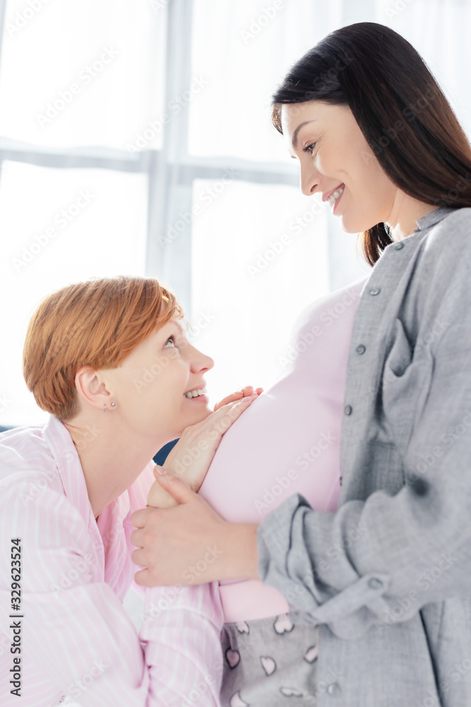 Side view smiling woman touching belly and looking at pregnant girlfriend in pajamas in bedroom Side view smiling woman touching belly and looking at pregnant girlfriend in pajamas in bedroom