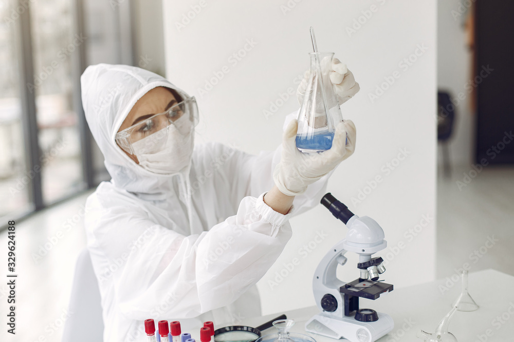A laboratory scientist is examining a blue solution in a bottle. The researcher is wearing a coverall suit and special glasses. A laboratory scientist is examining a blue solution in a bottle. The researcher is wearing a coverall suit and special glasses.