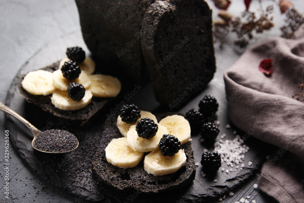 Healthy bread. Poppy seed black bread with banana and blackberries on a black board. Poppy seeds in spoon, salt, grey cloth. Background image, copy space
