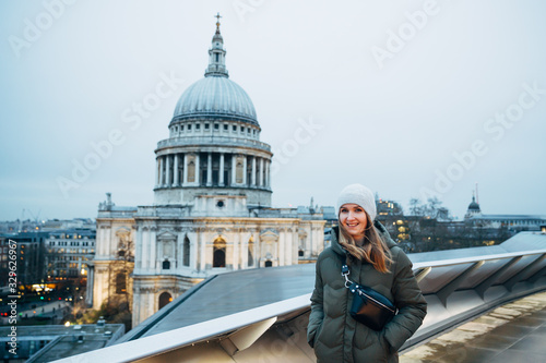 Pretty smiling tourist woman wearing winter hat, coat and trendy waist bag walking on the rooftop near St. Pauls Cathedral and enjoys evening cityscape. photo