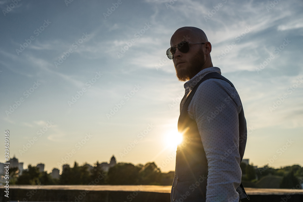 Bold Guy with a stylish beard and sunglasses on a blurred city background during sunset. Concept of success and will