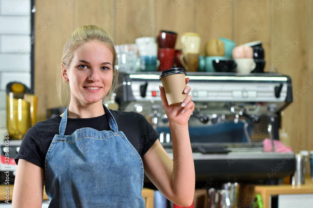 Obraz premium A young bartender girl in a jeans apron on a background of a coffee machine offers coffee to go. Concept of sale and work in a coffee shop in a small business.