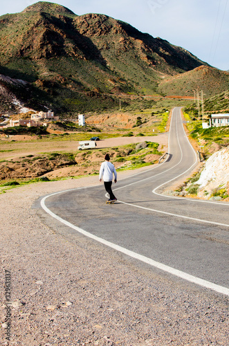 .young man with skateboard skating on a long road in a mountainous landscape