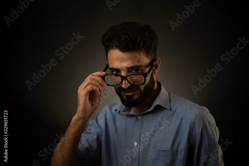 Portrait of young brunette Indian/European/Arabian/Kashmiri man in blue formal shirt and trousers with glasses in front of black copy space background. Indian lifestyle and fashion photography.