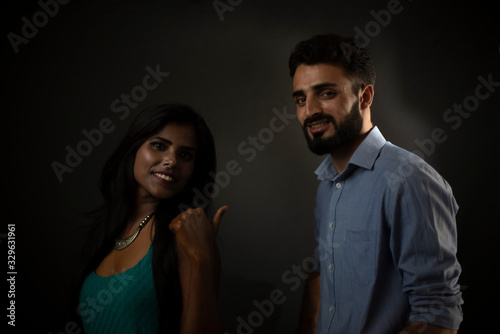 A dark skinned Indian/African girl and a Kasmiri/European/Arabian man in formal wear with shopping bag in front of a black copy space studio background. Indian lifestyle and fashion photography.