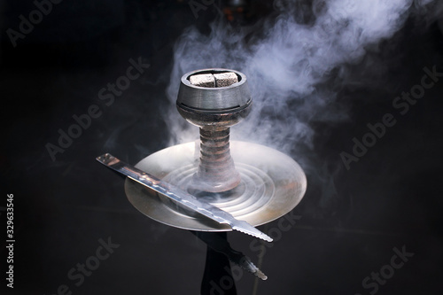 Bowl of hookah with kaloud and coals, a dish with tongs and smoke around