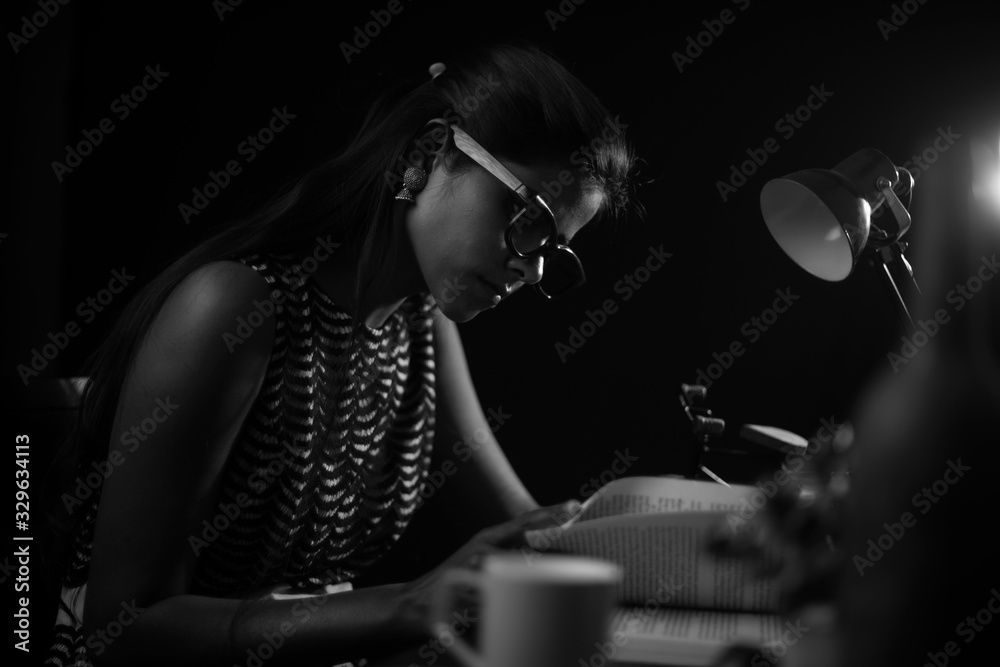 Young brunette Indian/African dark skinned lady in western dress and spectacles studying in front of a table lamp in black copy space studio background. Lifestyle and fashion.