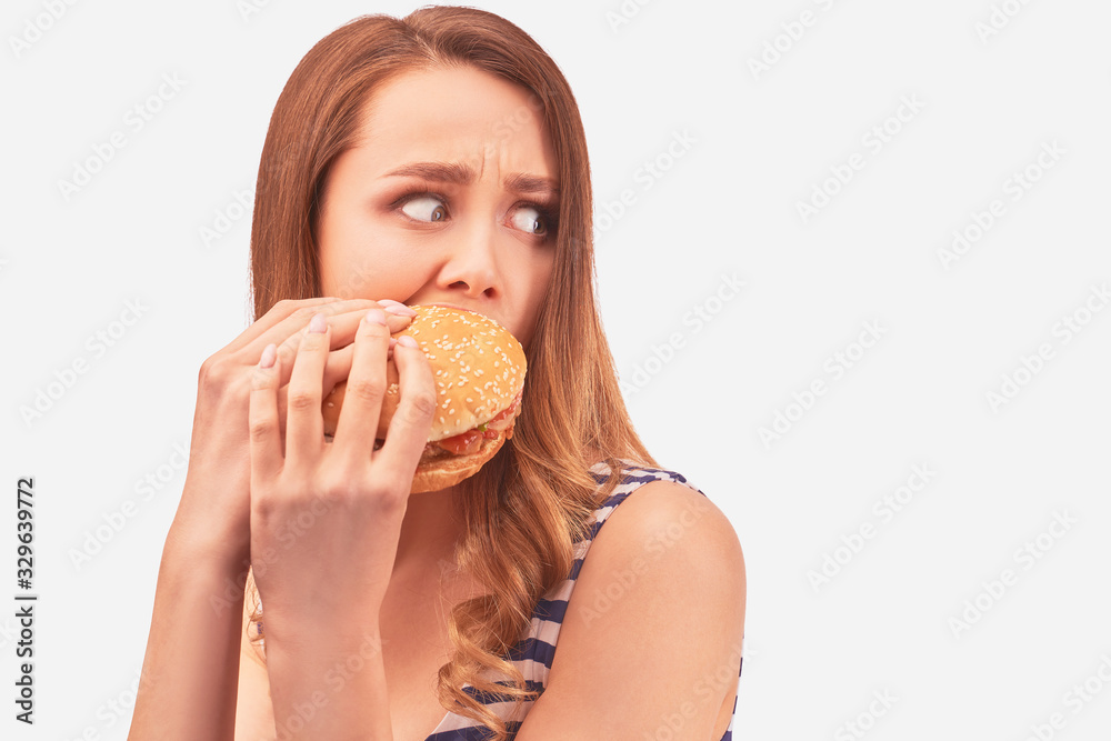 Cute girl quickly eats Burger, is afraid of being caught, hides from prying eyes. Wears striped t-shirt, isolated on white background in Studio with space for advertising Cute girl quickly eats Burger, is afraid of being caught, hides from prying eyes. Wears striped t-shirt, isolated on white background in Studio with space for advertising