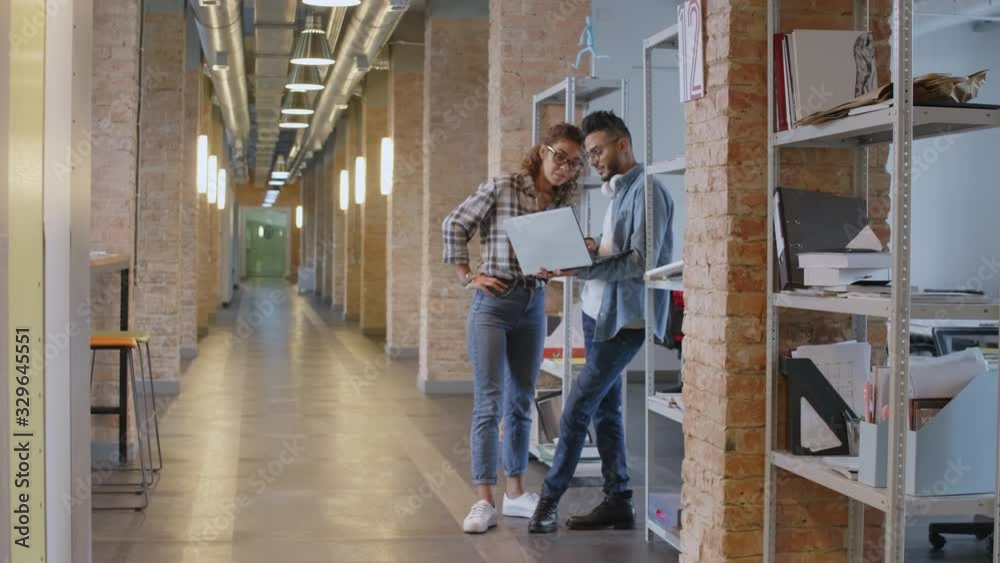 Young middle eastern man using laptop and speaking with mixed raced female colleague while standing together in loft hallway of modern office center Young middle eastern man using laptop and speaking with mixed raced female colleague while standing together in loft hallway of modern office center
